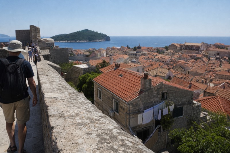 From high on Dubrovnik's city walls in Croatia, a walker's shoulder passes a worn limestone parapet, terracotta rooftops tumbling toward the sea, summer midday