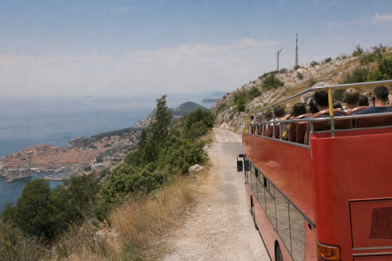 An open-top red panorama bus parked on the dusty upper road over Dubrovnik, Croatia, late morning haze, passengers seated high with headphones