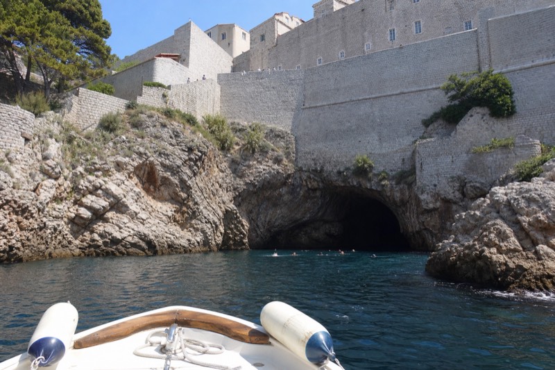 A small open boat noses toward the dark arched mouth of Betina Cave beneath Dubrovnik's sunlit city walls, sea chop glinting