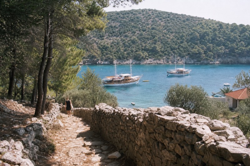 High on a rocky path above a swimming cove on Koločep, one of Croatia's Elaphiti Islands near Dubrovnik, midday sun flattens turquoise water where two wooden cruise boats wait