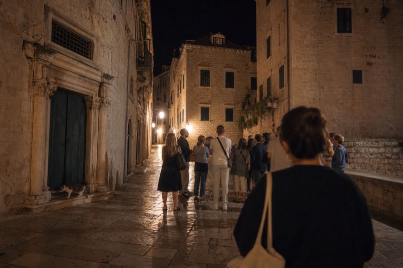 Night in Dubrovnik's Old Town, warm lamplight on wet polished limestone paving between medieval houses, a listener seen from behind at the edge of a small tour cluster