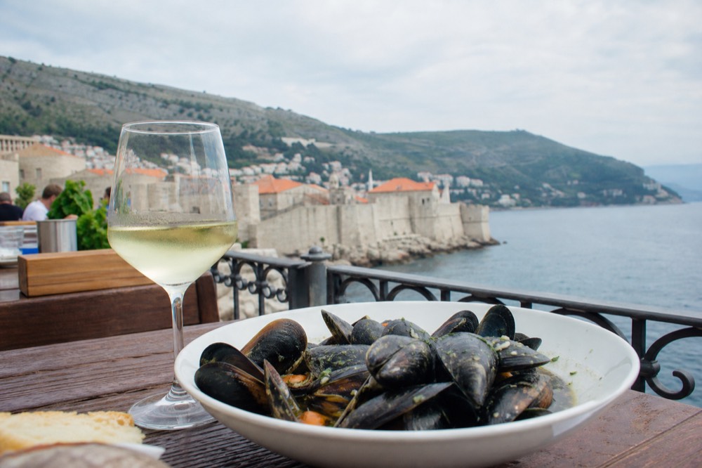 A bowl of fresh mussels in black broth beside a glass of white wine on a rustic restaurant table