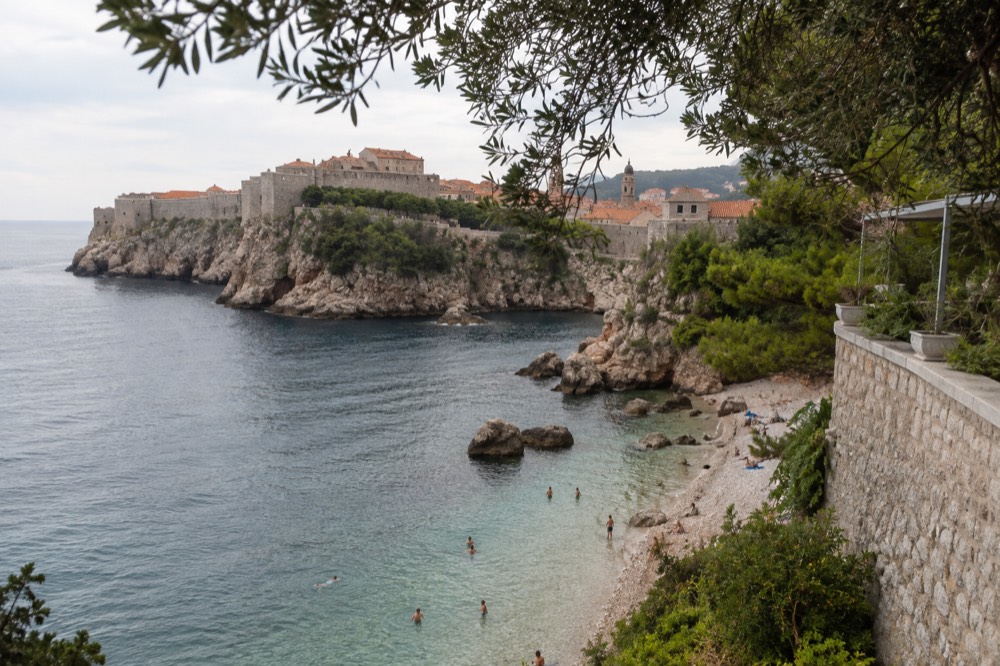 A pebble shoreline below the Old Town walls with swimmers in shallow turquoise water