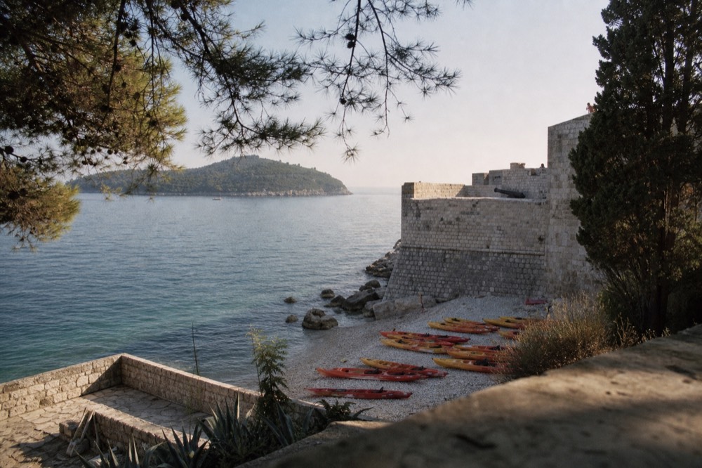 Sea kayaks lined up on a small pebble beach beneath stone city walls