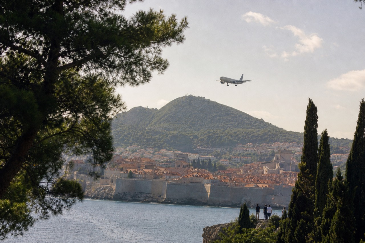 A plane descending over a walled Adriatic coastal town with terracotta rooftops and a green hillside behind it