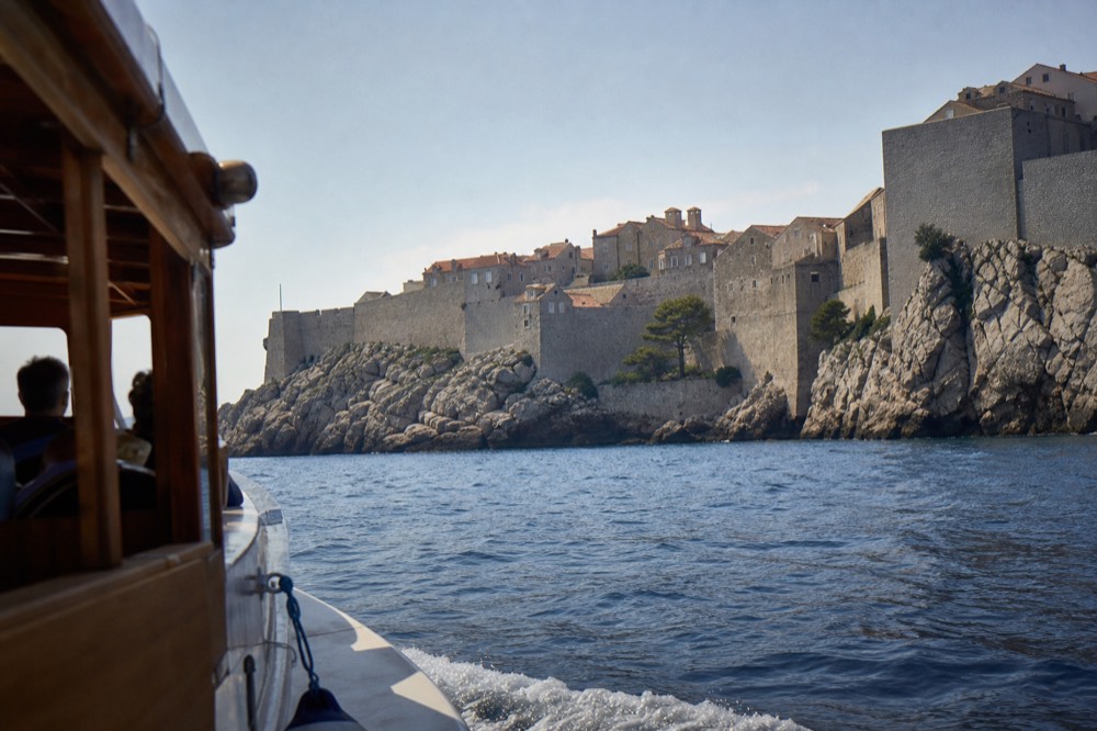 A wooden tour boat cruising past a walled Adriatic old town at midday