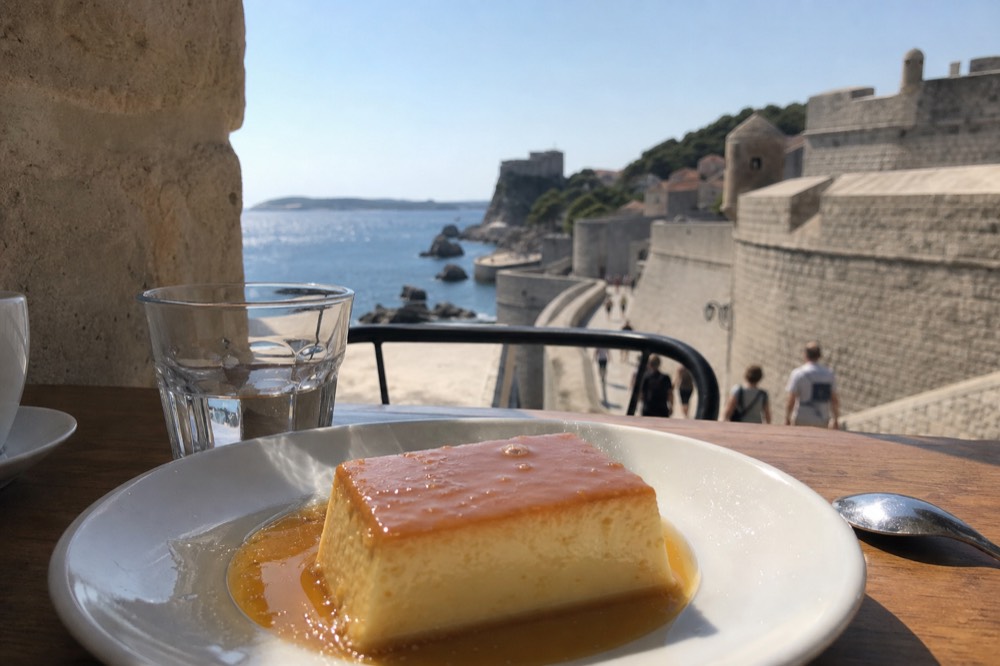 A close-up of a plate of rožata, caramel custard with a glazed top, on a restaurant table with a view of the Old Town walls