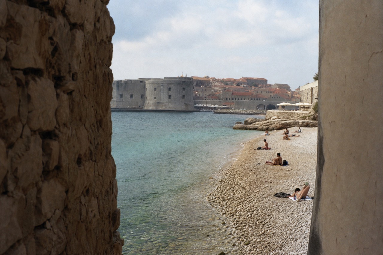 A calm pebble beach with clear shallow water and a walled old town in the background