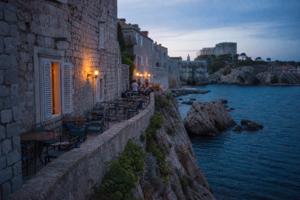 A simple terrace with tables and chairs on a rocky ledge above the sea below stone walls