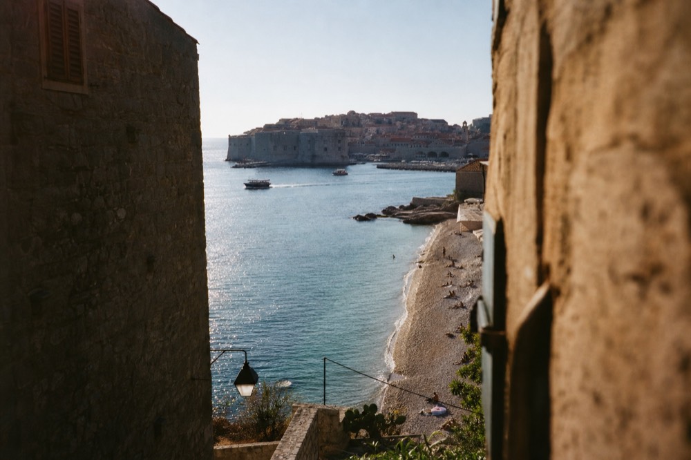 A crescent-shaped pebble beach with clear turquoise water and a view of the Old Town in the distance