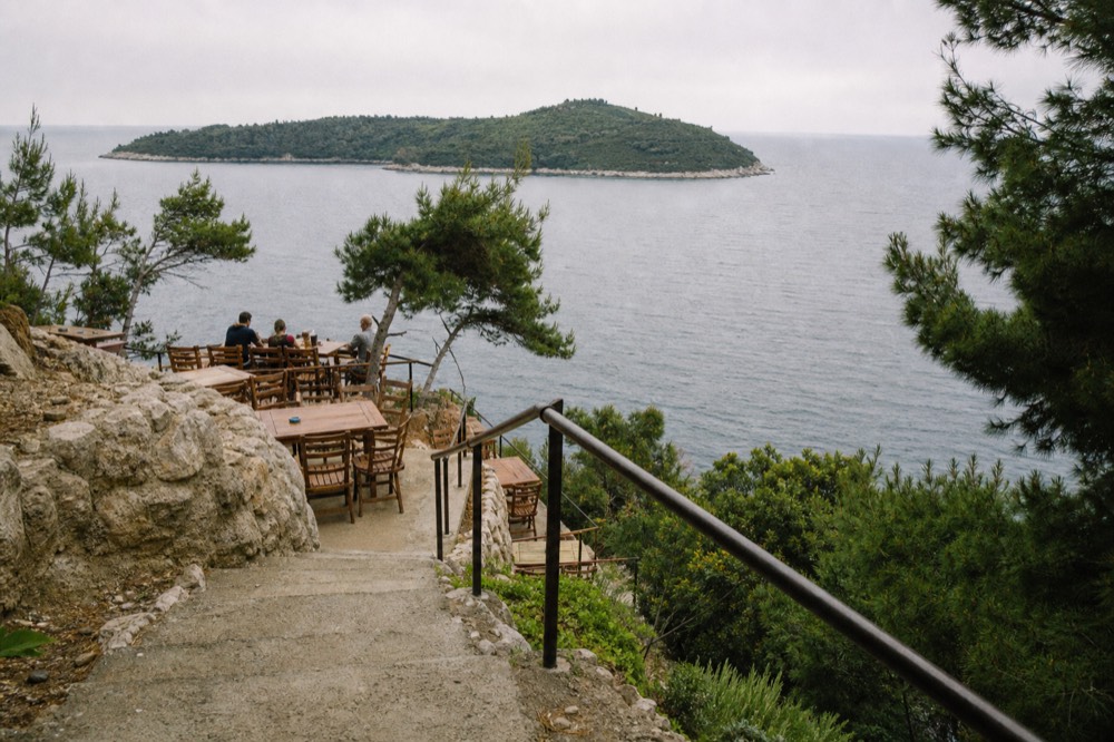 Wooden tables and chairs on a rocky ledge above the sea with the island of Lokrum in the distance