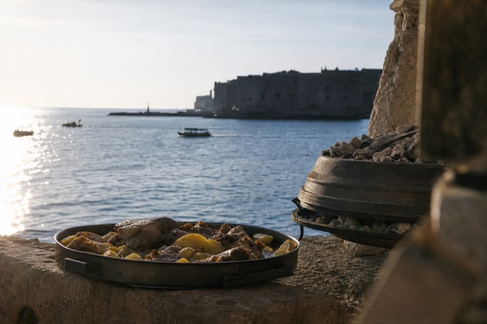 An iron peka dome resting on coals beside a tray of lamb and potatoes
