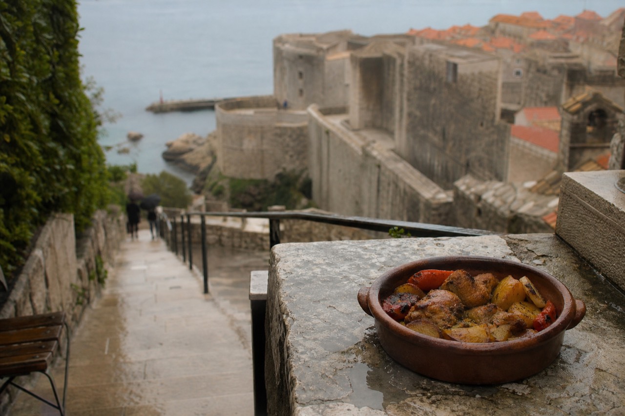 A clay dish of slow-cooked meat and vegetables resting on a stone terrace table beside the Dubrovnik city walls