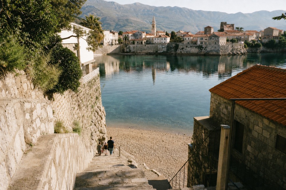 Stone houses and a church tower reflected in still bay water with mountains rising behind