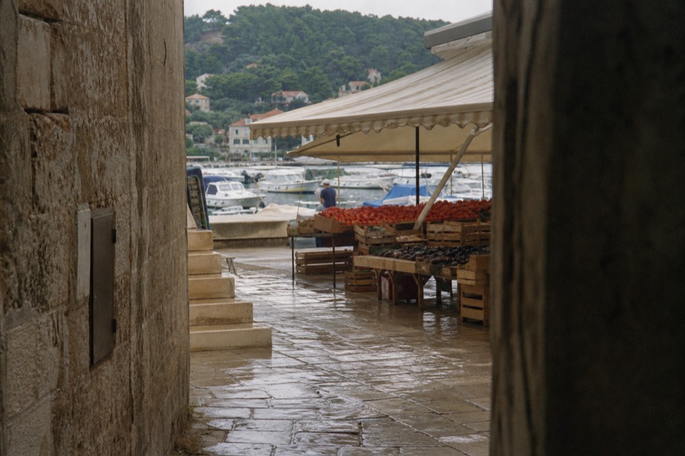 A market stall piled with fresh figs and tomatoes under an awning by the harbour