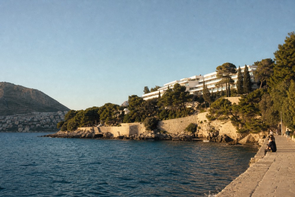 A white hotel building with terraces set among pine trees above a bay