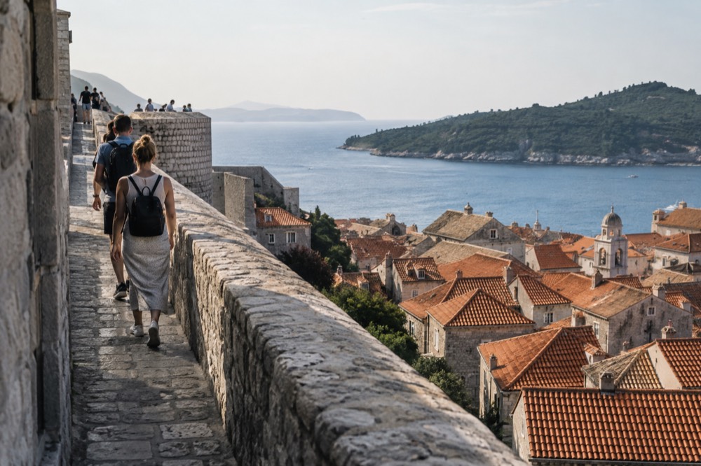 Visitors walking the top of a stone city wall above red rooftops and the sea