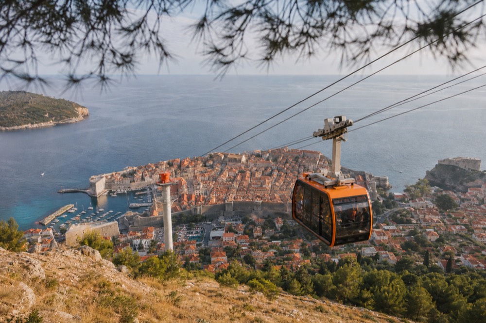 A cable car cabin ascending above red rooftops toward a hilltop station in late afternoon light