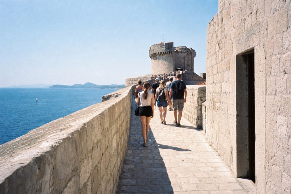 A crowd of visitors walking on the city walls near the Minceta Tower, with the sea visible on one side