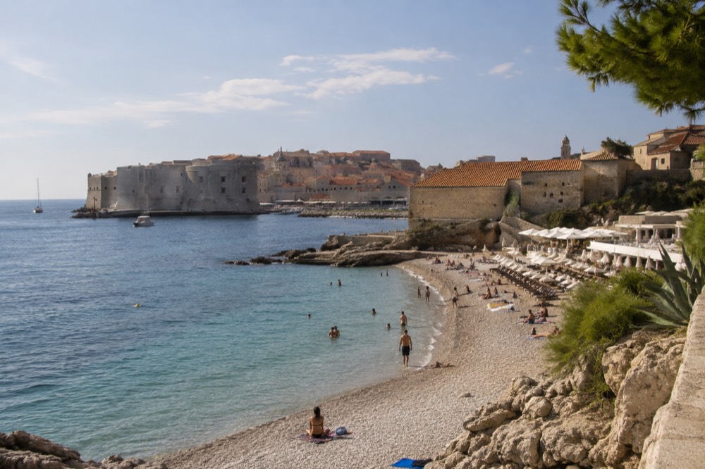 A narrow pebble beach between clear water and stone buildings, a few swimmers in the shallows