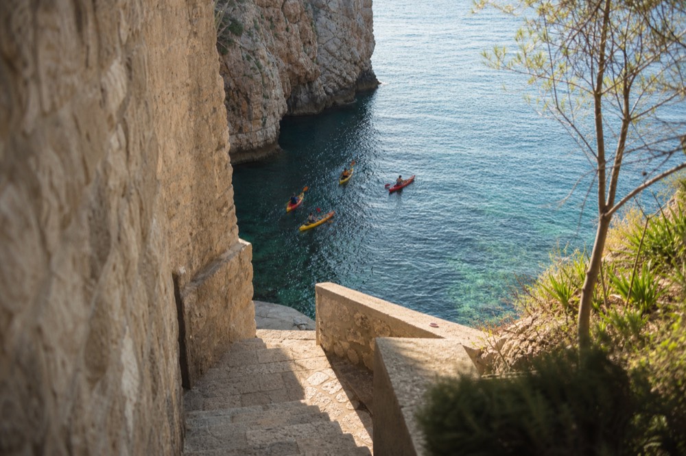 A line of sea kayaks in bright water below steep stone walls, paddlers close to the cliffs