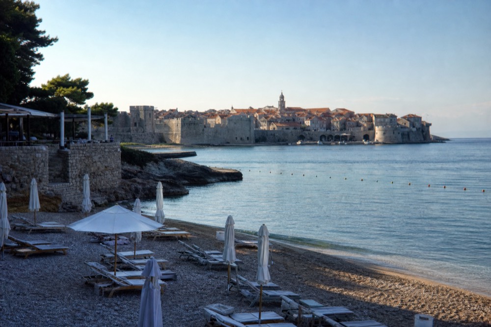 An old town with orange roofs and a bell tower seen across calm water in early light