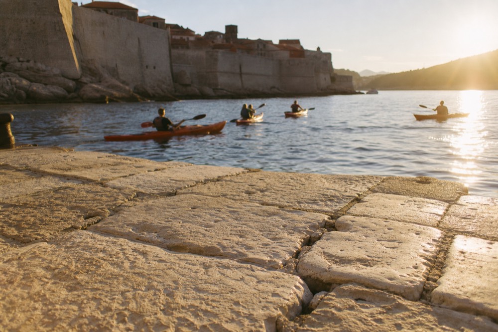 Sea kayaks on calm water at sunset below a walled city