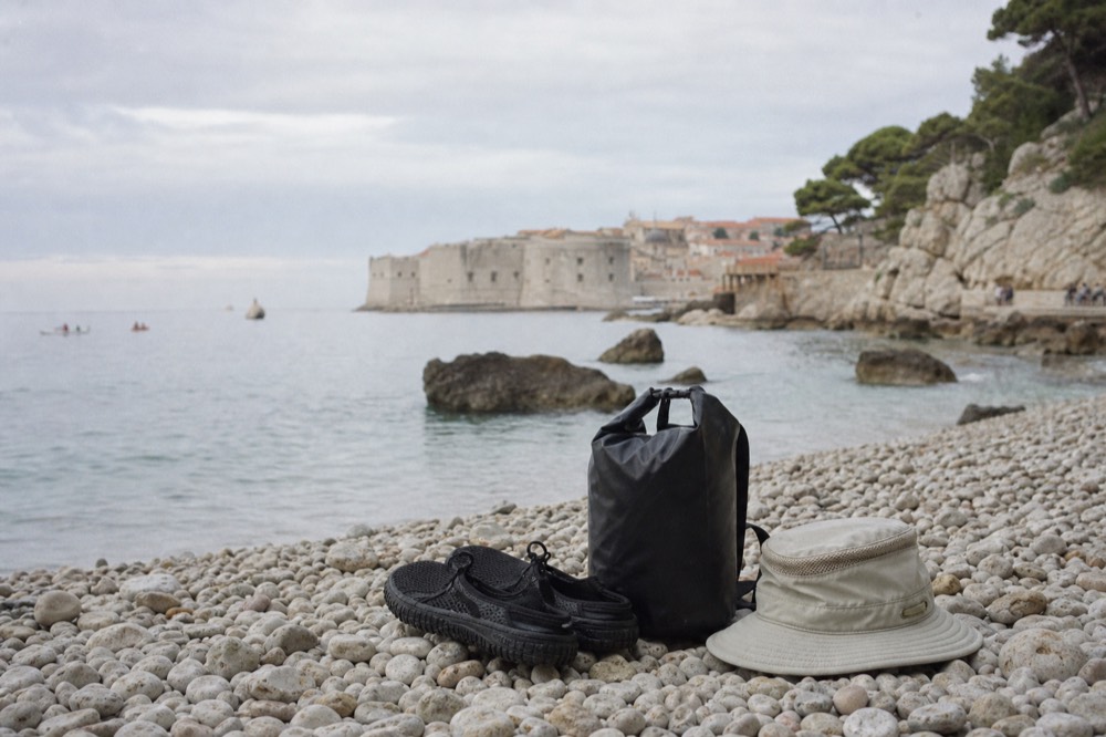 Water shoes, a hat and a dry bag arranged on smooth pebbles beside the sea