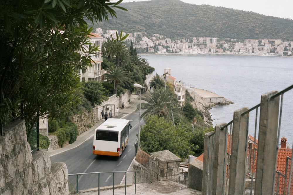 A local bus pulling away from a stop with apartment buildings and palm trees along the street