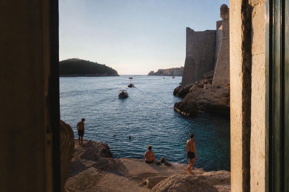 People swimming from flat rocks into clear blue water beneath high stone walls