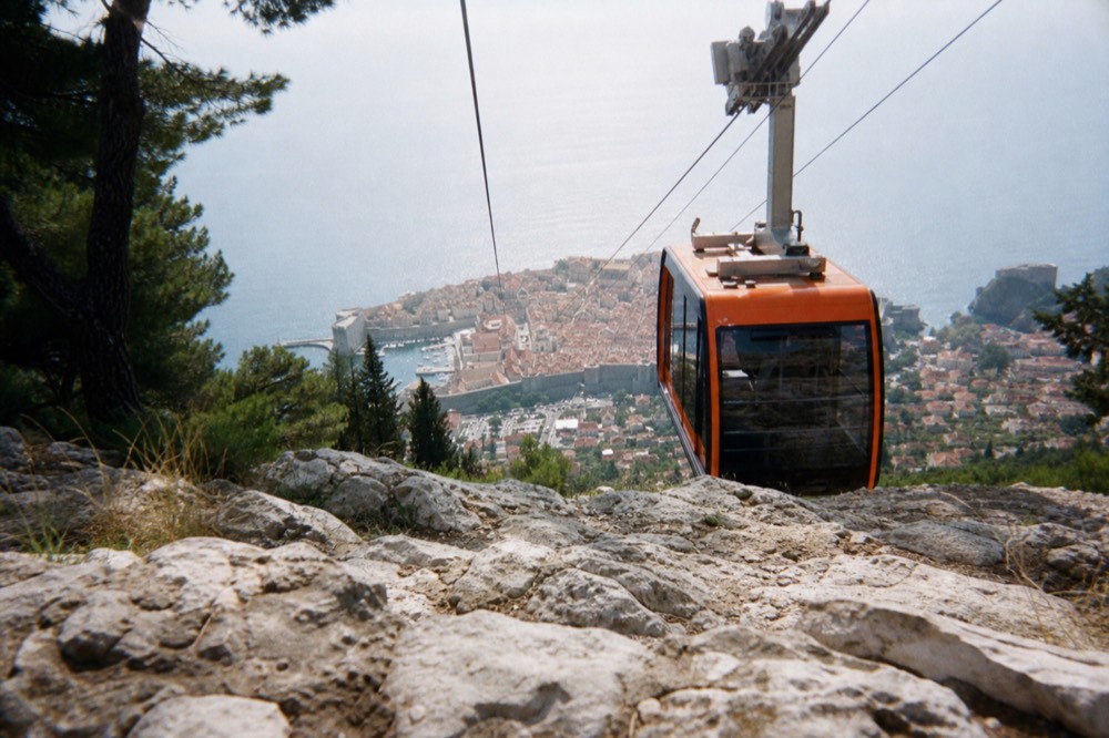 The orange Dubrovnik cable car cabin ascending over the red-roofed Old Town with the Adriatic Sea in the background