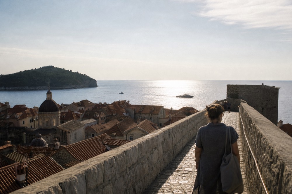 A walker on the Dubrovnik city walls looking down over red rooftops and the sea