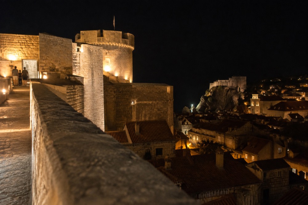 A stone fortress terrace at night lit by warm lights above dark rooftops