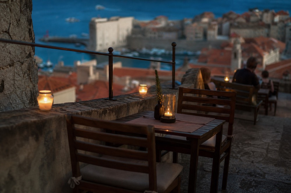 A stone terrace at night with small tables, candles and a view over city rooftops