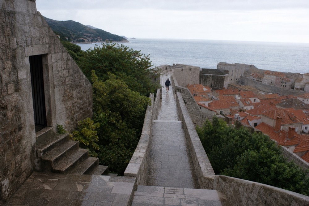 A stone city wall walkway above red rooftops with the sea beyond