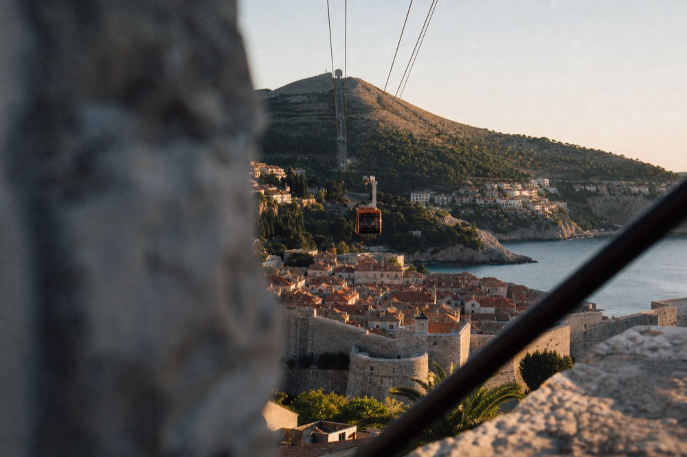 A cable car cabin ascending above a walled city toward a hilltop station at dusk