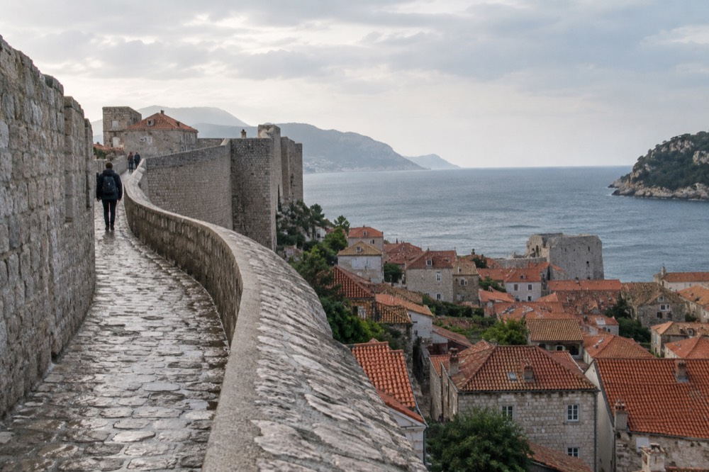 A stone city wall walkway curving above red rooftops and the sea
