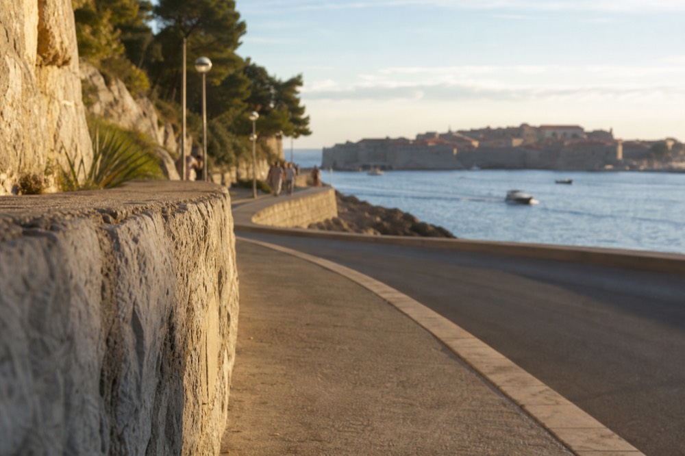 Coastal road and pavement curving along a bay toward Dubrovnik's walled city