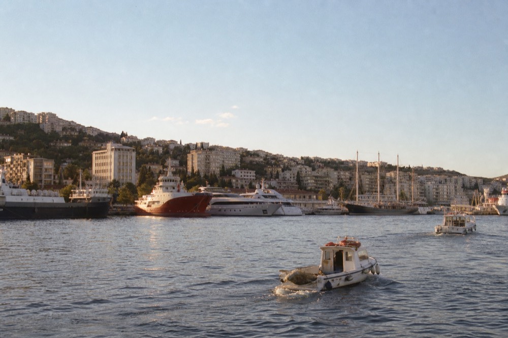 A wide harbour with moored ships, apartment blocks and hills behind