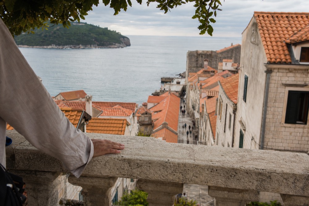 A guide's hand resting on a sun-warmed stone balustrade above a narrow Old Town street