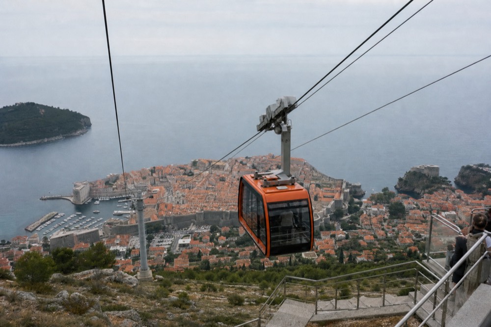 A red cable car cabin ascending above Dubrovnik's terrac rooftops toward a hilltop station