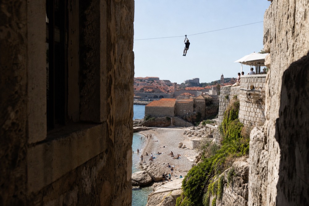 A zipline rider in a harness crossing a cable high above Dubrovnik's red rooftops