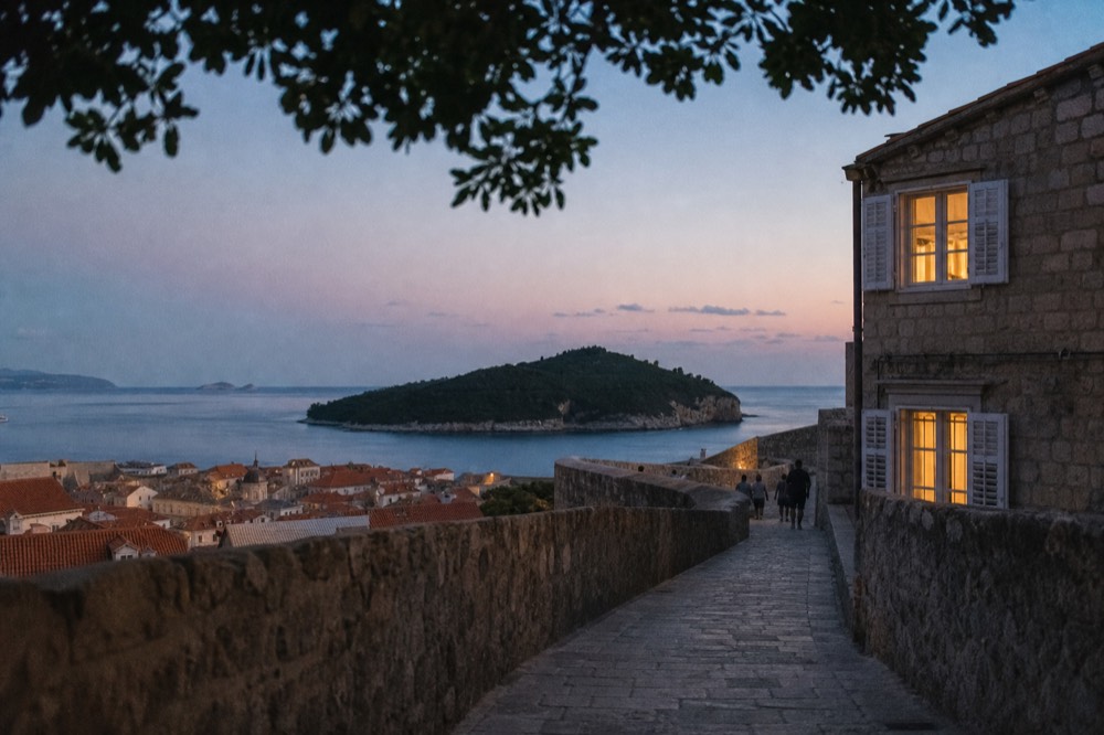 A wide stone walkway on a fortress wall curving above rooftops and a green island at sunset