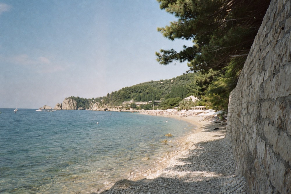 A pebble beach with clear shallow water, sun umbrellas and wooded hills behind