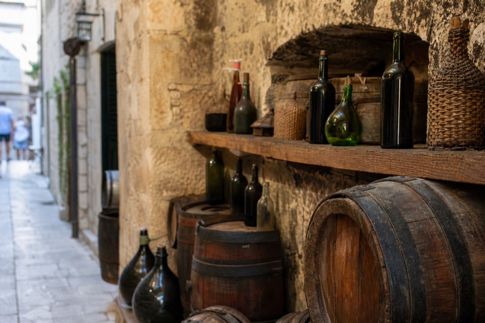 A rustic stone cellar with wooden barrels and bottles lined along a shelf
