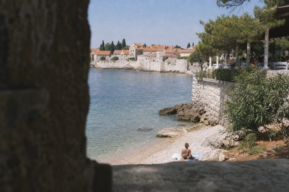 A pebble beach with clear water and a walled old town in the background