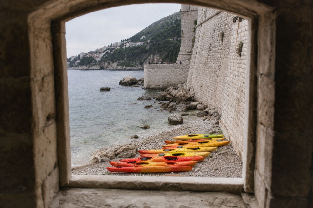 Sea kayaks lined up on a rocky shore beside clear water near a stone wall