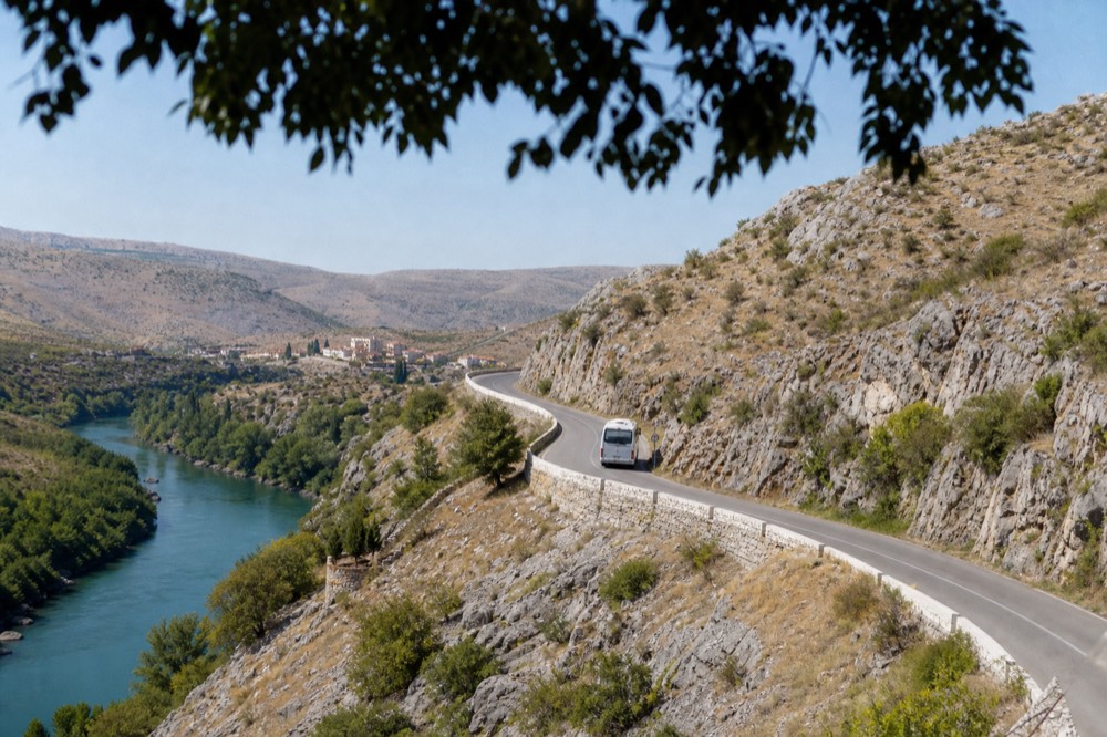 A tour minibus on a winding road through dry rocky hills above a river valley