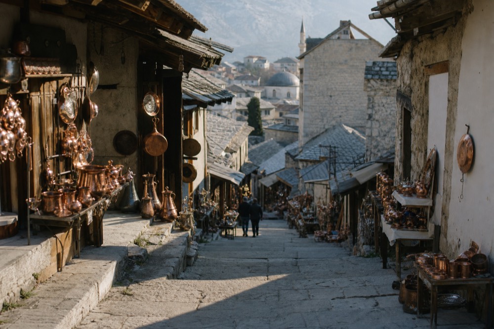 A narrow bazaar street lined with copper shops and stone buildings under a bright sky