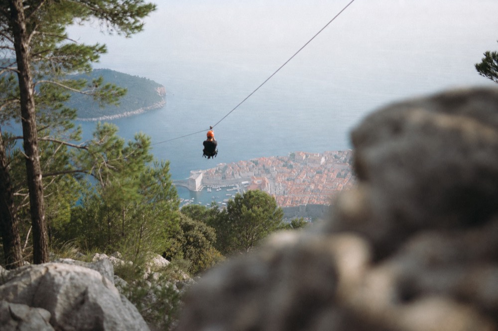 A person on a zipline high above the hillside with Dubrovnik and the sea far below